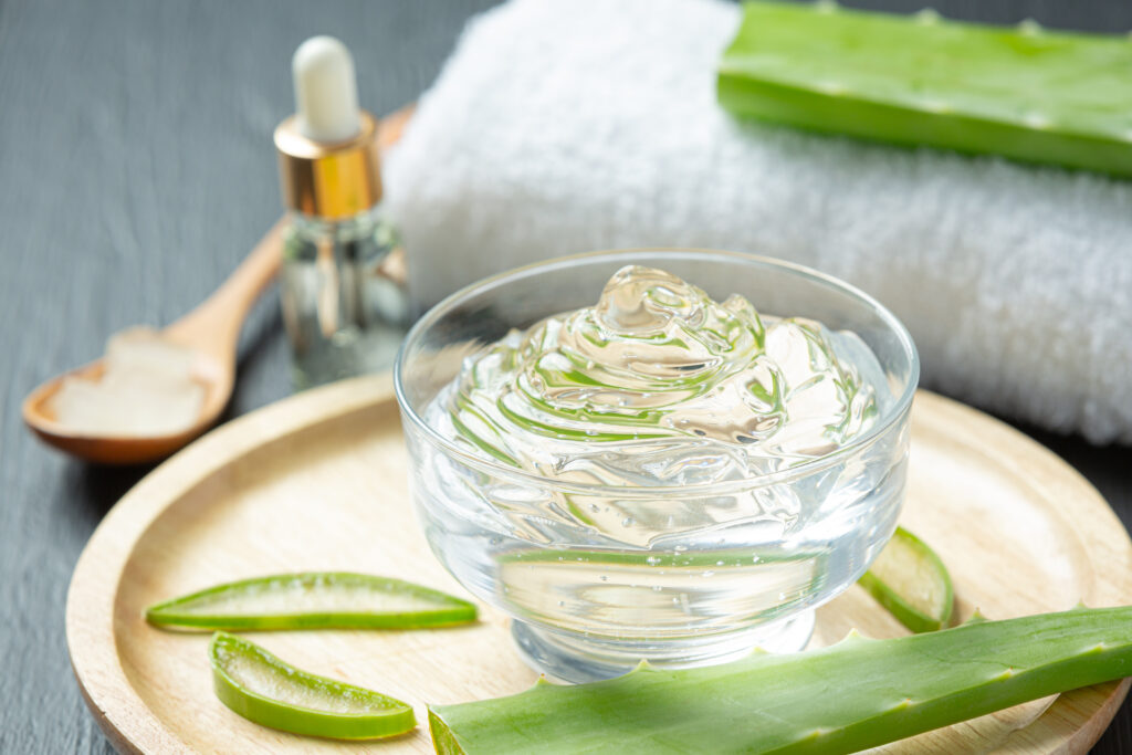 Un tazón de vidrio lleno de gel de aloe vera transparente en un set de spa natural con una bandeja de madera, suero facial, y una toalla blanca de fondo.