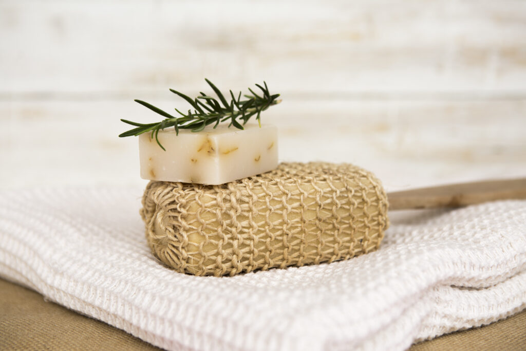 Mano cubierta de espuma blanca sosteniendo una barra de champú sólido redonda y amarilla con una rodaja de cítrico seco incrustada, bajo una luz contrastada que crea una sombra marcada.