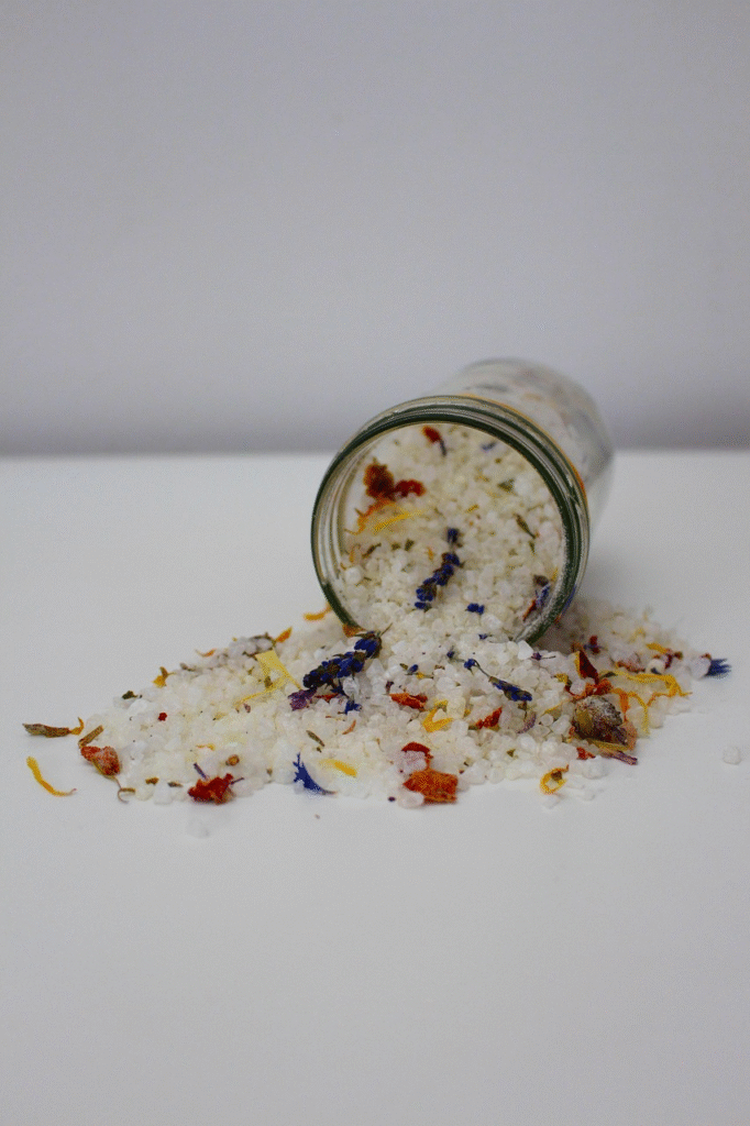 Frasco de vidrio volcado derramando sales de baño blancas mezcladas con flores secas de lavanda, caléndula y hierbas, ingredientes naturales para recuperación muscular