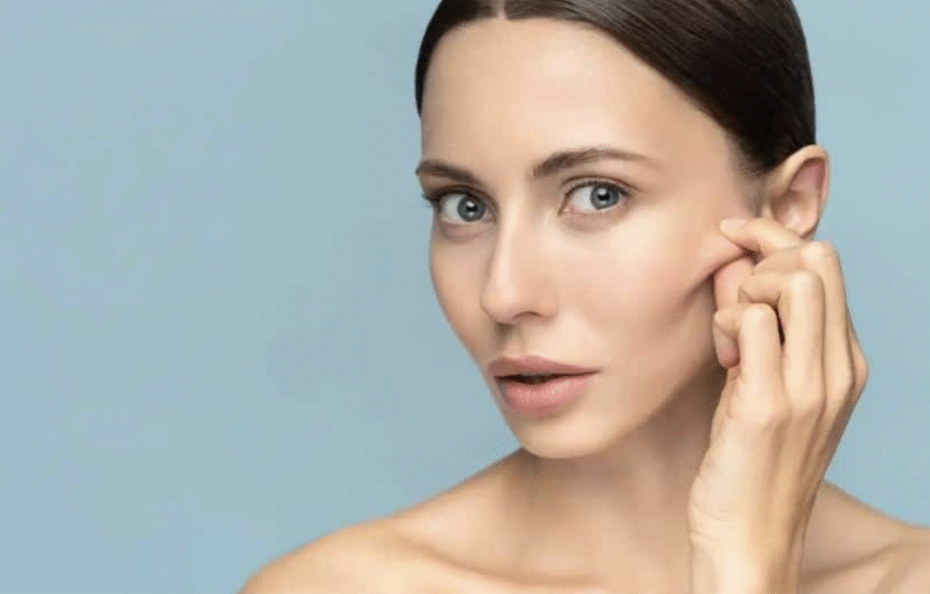 Mujer joven pellizcando suavemente la piel de su mejilla para mostrar elasticidad y firmeza sobre un fondo azul claro.