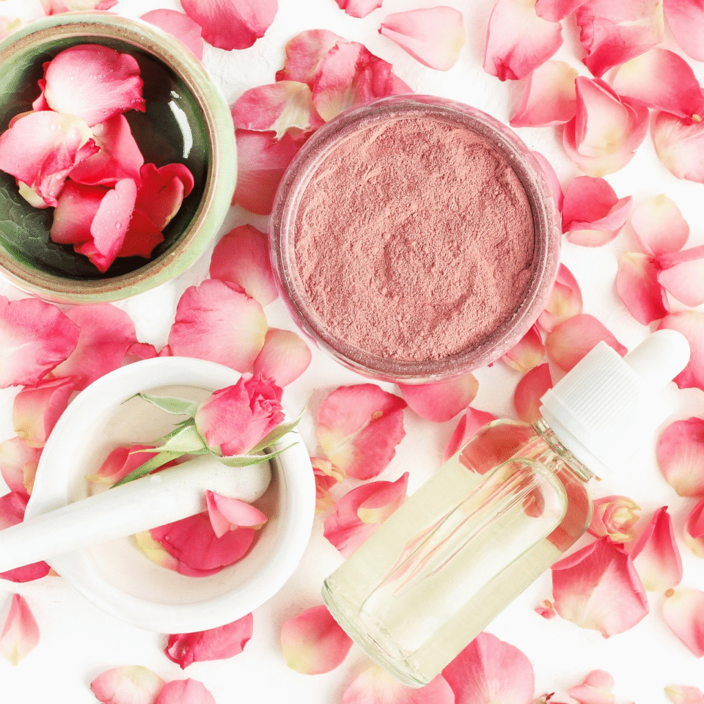 Vista superior de un set de belleza natural que incluye crema o polvo de arcilla rosa en frasco, un mortero con una rosa fresca, un gotero con aceite esencial y pétalos dispersos.