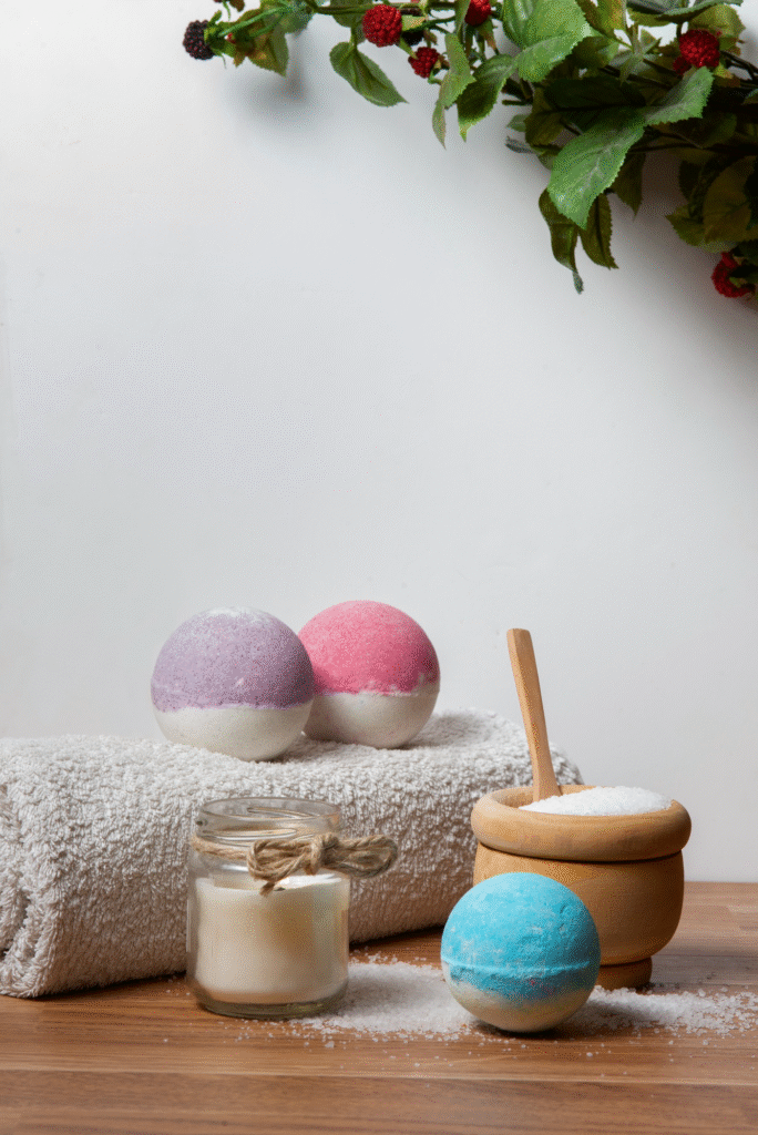 Bombas de baño efervescentes de colores y sales de mar naturales en mortero de madera junto a una vela aromática y toalla blanca, ideal para un spa relajante en casa.