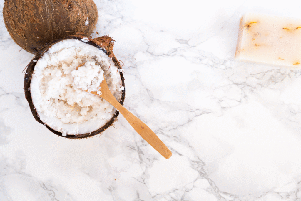 Exfoliante natural de coco y azúcar dentro de un coco fresco con cuchara de madera y jabón artesanal sobre fondo de mármol