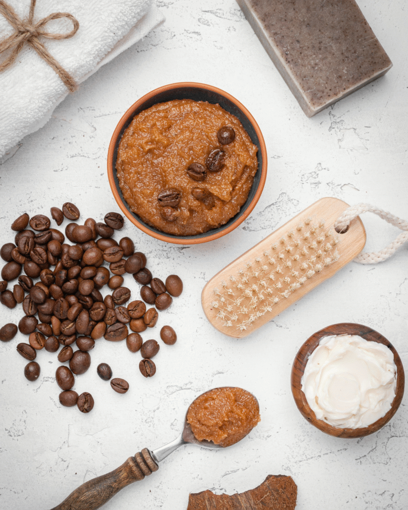 Composición de productos de spa naturales sobre fondo blanco, incluyendo un cuenco de exfoliante de café con granos enteros, granos de café sueltos, un cepillo de madera, un cuenco de crema blanca, una barra de jabón artesanal, una toalla y una cuchara con exfoliante.