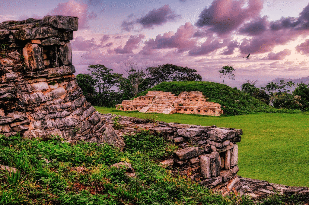 Ruinas arqueológicas de piedra con nichos bajo un cielo de atardecer en tonos púrpura y rosa, rodeadas de vegetación y césped verde en México.