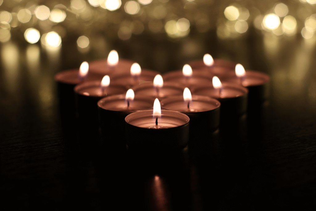 Velas de té encendidas dispuestas en forma de corazón sobre una superficie oscura con un fondo de luces bokeh doradas y cálidas.