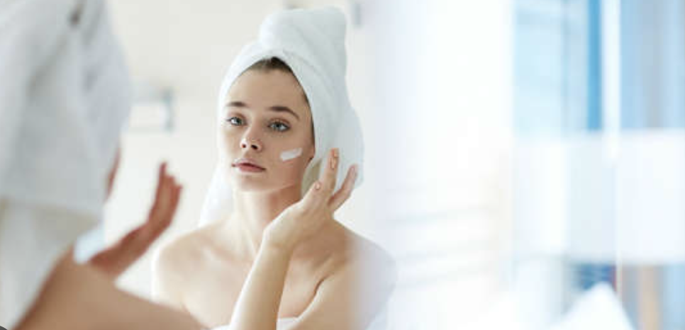 Mujer joven con una toalla blanca en la cabeza aplicándose una mancha de crema hidratante en la mejilla mientras se mira en el espejo del baño.