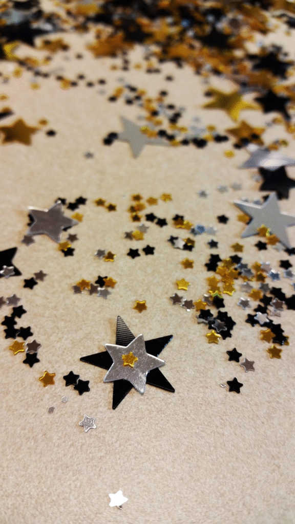 Mix de glitter biodegradable con forma de estrellas negras, plateadas y doradas de Piñuela esparcidas sobre fondo neutro