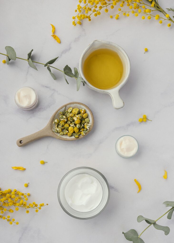 Frasco gotero blanco minimalista rodeado de flores frescas de manzanilla y un cuenco con mezcla de crema de manzanilla piel y aceite bajo luz solar directa.